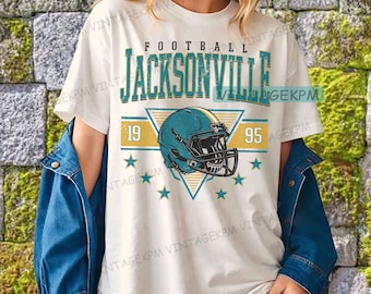 May include: Cream-coloured t-shirt with a teal and gold graphic. The design includes the word "FOOTBALL" above "JACKSONVILLE" in teal, a teal American football helmet, the numbers "19" and "95", and teal stars. A blue denim jacket is worn.