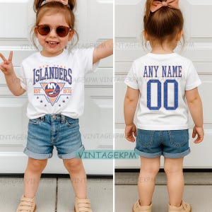May include: A white t-shirt featuring the "New York Islanders" logo and "EST. 1972" text. The shirt is worn with denim shorts. The back of the shirt displays "ANY NAME" and the number "00" in blue.