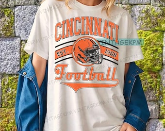 Può includere: T-shirt color crema con la scritta "Cincinnati Football" in arancione, insieme a una grafica di casco da football. La maglietta presenta anche il testo "EST. 1968". Indossata con una giacca di jeans blu.