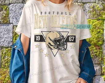 May include: Cream-colored t-shirt with a football-themed graphic. The design features the words "FOOTBALL NEW ORLEANS" above a football helmet inside a triangle, with the numbers "19" and "67" on either side, and stars below.