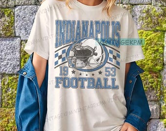 May include: White t-shirt with a blue and white graphic that says "Indianapolis Football 1953". The graphic includes a football helmet and stars. The shirt has short sleeves and a crew neck.