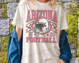 Op de afbeelding: Wit T-shirt met een verweerde roze opdruk met de tekst "ARIZONA FOOTBALL" en een illustratie van een footballhelm. Het shirt heeft ook de nummers "19" en "20" en sterren. De persoon draagt een blauw spijkerjasje.