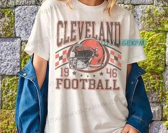 May include: Off-white t-shirt with a vintage-style graphic of a football helmet and the words "Cleveland Football 1946." The design incorporates red, black, and white colours. The shirt is worn by a person with a denim jacket.