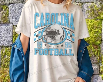 Może przedstawiać: Kremowa koszulka z motywem futbolu amerykańskiego Carolina. Projekt zawiera napis "CAROLINA" w jasnoniebieskim kolorze, hełm futbolowy, liczby "19" i "95" oraz słowo "FOOTBALL". Koszulka ma styl vintage.