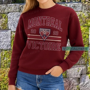 May include: A maroon sweatshirt with the word "MONTREAL" arched across the top in white lettering. Below, the word "VICTOIRE" is displayed. A crest with a shield and the text "20 VICTOIRE 23" is centered. The sweatshirt has long sleeves.