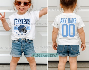 Può includere: Maglietta bianca con "TENNESSEE" in blu, una grafica di casco da football e i numeri "19" e "60". La parte posteriore della maglietta ha "ANY NAME" e il numero "00" in blu. Indossata con pantaloncini di jeans.