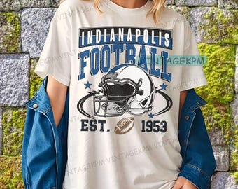 May include: White t-shirt featuring a blue and black graphic. The design includes the words "Indianapolis Football" above a football helmet and football, with "EST. 1953" below. The shirt is a casual style.