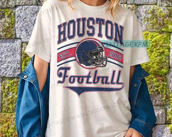 Op de afbeelding: Crèmekleurig T-shirt met een verweerde afbeelding. Het marineblauwe en rode ontwerp toont "HOUSTON Football", een footballhelm en het jaar "2002". Het shirt wordt gedragen met een blauw spijkerjasje.