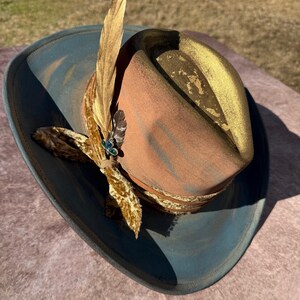 May include: A brown and blue cowboy hat with a gold trim and a feather accent. The hat has a velvet ribbon with a jeweled pin.