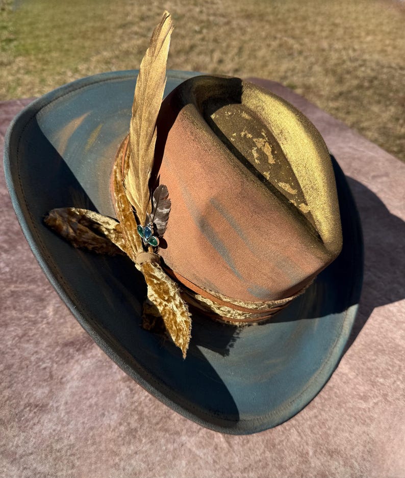 May include: A brown and blue cowboy hat with a gold trim and a feather accent. The hat has a velvet ribbon with a jeweled pin.