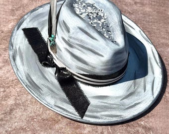 May include: A silver fedora hat with a black ribbon and a white stripe. The hat has a textured surface and a small feather accent.