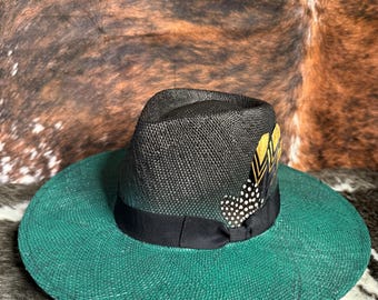 May include: A fedora-style hat with a wide, emerald green brim and a black crown. A black ribbon encircles the base of the crown. The hat is adorned with a decorative feather with gold and white accents.