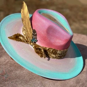 May include: A pink and blue ombre cowboy hat with a gold feather and a velvet ribbon. The hat has a wide brim and a tall crown.