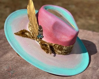 May include: A pink and blue ombre cowboy hat with a gold feather and a velvet ribbon. The hat has a wide brim and a tall crown.