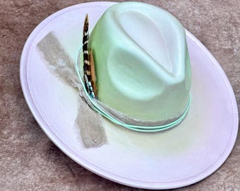 May include: A light green fedora hat with a wide brim. The hat features a decorative feather and a light green band around the crown. The brim is a light pink color with a faded design.