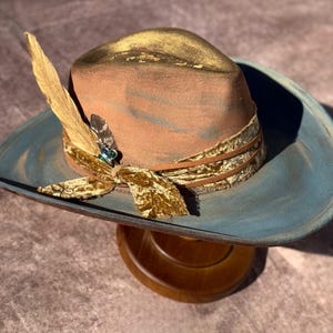May include: A brown and blue cowboy hat with a gold trim and a feather accent. The hat has a velvet band with a gold pattern.