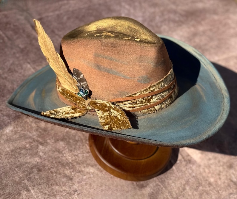 May include: A brown and blue cowboy hat with a gold trim and a feather accent. The hat has a velvet band with a gold pattern.