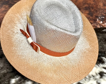 May include: A stylish fedora hat with a silver crown and a wide, tan brim. The hat features a rust-colored band and is adorned with a decorative feather and ribbon. The hat is a fashionable accessory.