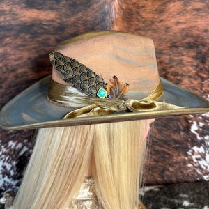 May include: A wide-brimmed hat with a brown and gray patterned crown and a gold velvet band. The hat is decorated with a gold and black feather and a turquoise gemstone.
