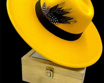 May include: A bright yellow fedora hat with a black band and a feather accent. The hat is sitting on a wooden box with a brass latch.