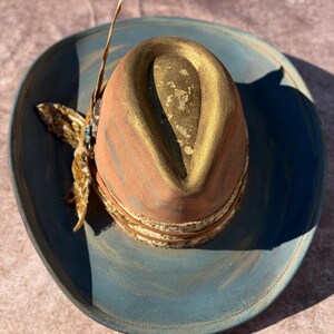 May include: A blue and gold cowboy hat with a brown leather band and a feather accent. The hat has a distressed look and is decorated with gold paint.