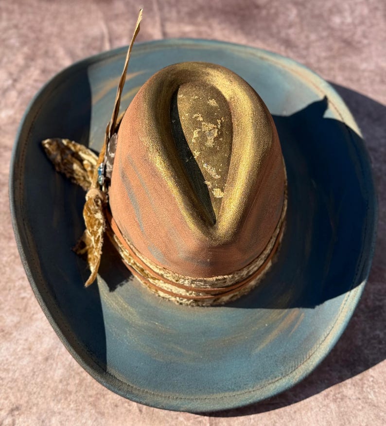May include: A blue and gold cowboy hat with a brown leather band and a feather accent. The hat has a distressed look and is decorated with gold paint.