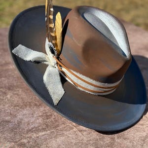 May include: A brown and gray fedora hat with a white ribbon and a feather accent. The hat has a distressed look and is made of a soft material.