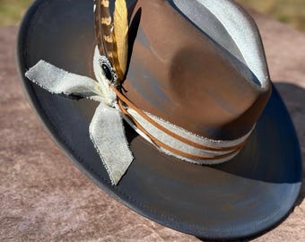 May include: A brown and gray fedora hat with a white ribbon and a feather accent. The hat has a distressed look and is made of a soft material.