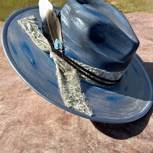 May include: A blue and grey cowboy hat with a grey velvet band and a feather accent. The hat has a distressed look and is decorated with a turquoise stone.