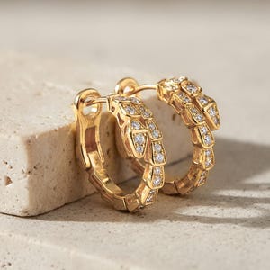 Puede incluir: Un par de pendientes de aro dorados con un diseño de serpiente. Los pendientes están incrustados con pequeñas piedras preciosas transparentes. Se muestran sobre una superficie clara y texturizada.