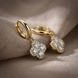 Peut inclure: Une paire de boucles d'oreilles créoles dorées avec un pendentif en forme de trèfle à quatre feuilles. Chaque pendentif est orné de petites pierres rondes transparentes disposées en motif floral, avec une pierre centrale entourée de pavés.