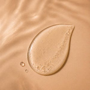 May include: A close-up of a clear, viscous liquid in a teardrop shape, with small bubbles and two smaller droplets. The liquid is light amber in colour, set against a beige background with subtle, rippled textures.