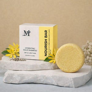 May include: A solid shampoo bar and its box are displayed on a white stone surface. The box is white with a yellow stripe and the text "NOURISH BAR". The bar is round and yellow. The product is for dry and curly hair.