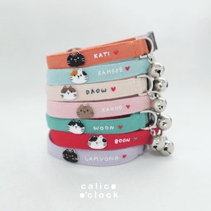 Puede incluir: Una pila de siete collares de tela para mascotas en naranja, azul claro, crema, rosa, verde azulado, rojo y lavanda. Cada collar presenta una cara de gato bordada, un nombre, un pequeño corazón rojo y un cascabel de metal plateado con hebilla.