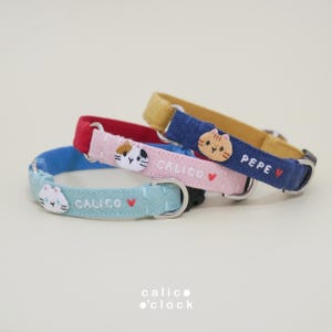 Puede incluir: Tres collares de tela para mascotas con caras de gato y nombres bordados. El collar azul claro dice CALICO con un corazón rojo, el rosa dice CALICO con un corazón rojo y el azul oscuro dice PEPE con un corazón rojo y borde amarillo.
