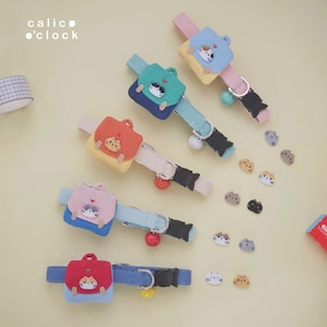 May include: A selection of cat collars featuring backpack-shaped charms in various colours, including red, blue, pink, and green. Each charm displays a different cat face design. The collars are fitted with bells and black buckles. The brand name "calic o'clock" is visible.
