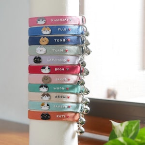 May include: A stack of 10 colorful cat collars with embroidered names and cat faces. Each collar has a small bell attached.