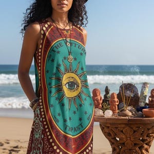 Puede incluir: Vestido sin mangas con un diseño celestial que presenta un sol con un ojo, rodeado de estrellas y las palabras "THE FUTURE" y "THE PAST". El vestido es en tonos de verde azulado, rojo y dorado. Un sombrero negro y joyas completan el look.