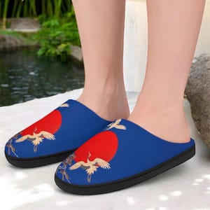 May include: Blue slippers with a red sun and crane design. The slippers have a black sole and a soft, comfortable interior. The design features a red sun and white cranes, creating a serene and artistic look.