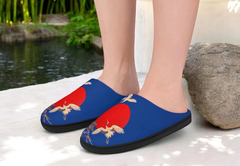 May include: Blue slippers with a red sun and crane design. The slippers have a black sole and a soft, comfortable interior. The design features a red sun and white cranes, creating a serene and artistic look.