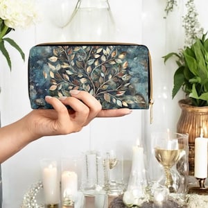 May include: A rectangular wallet with a nature-inspired design. The wallet features a detailed illustration of a tree with leaves in shades of brown, green, and gold against a blue background. It has a gold zip and is held by a person.