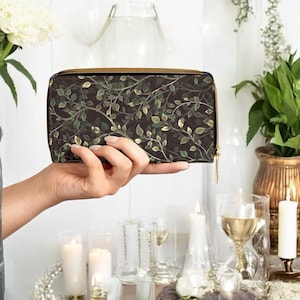 May include: A rectangular wallet with a dark background and a pattern of green leaves and vines. The wallet has a gold zipper and is held in a hand. The background includes candles, flowers, and glassware.