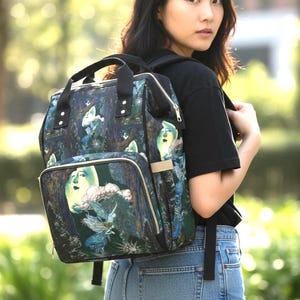 May include: A backpack with a surreal design featuring faces, clouds, and fairies in blue, green, and white. It has a top handle, a front pocket, and adjustable straps. The bag is a dark colour with a zip.