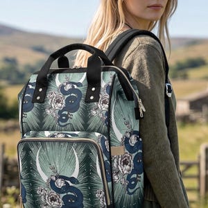 May include: A backpack featuring a dark green background with a repeating pattern of black snakes, white peonies, and white crescent moons. The bag has black top handles, black shoulder straps, and a front zippered pocket with metallic zips.