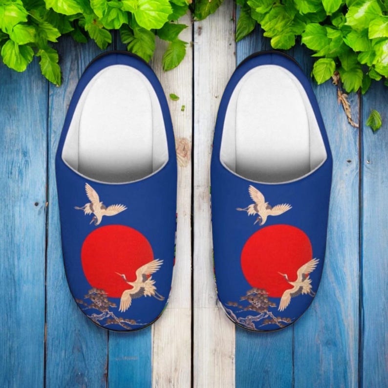 May include: A pair of blue slippers with a Japanese-inspired design. Each slipper features a red sun, flying cranes, and a landscape scene. The interior is white.