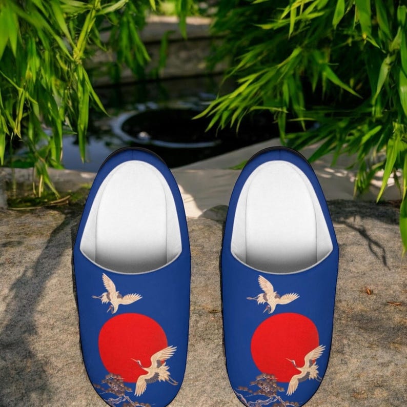 May include: A pair of blue slippers with a white interior. Each slipper features a red sun design with white cranes and a detailed wave pattern. The slippers are designed for indoor use.