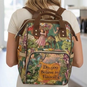 May include: A backpack featuring a colorful fairy and mushroom forest print with brown straps and trim. The front pocket displays the text Do you Believe in Fairies? in a decorative font. The bag includes a top handle and adjustable shoulder straps.