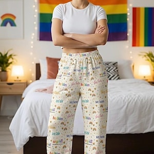 Puede incluir: Pantalones de pijama color crema con gráficos de arcoíris y pronombres. Los pantalones presentan los pronombres "he/him", "she/her", "they/them" y "ze/zir". Se usa una camiseta blanca con los pantalones. Una bandera arcoíris cuelga en el fondo.