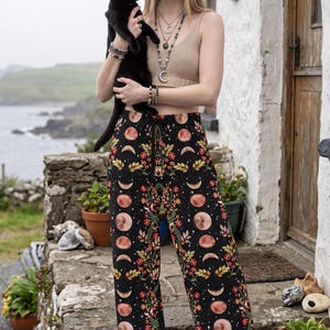 May include: A beige ribbed crop top and black wide-leg trousers featuring a repeating pattern of moon phases, colourful flowers, and green leaves. The trousers have a drawstring waist and a loose fit, paired with layered necklaces and a black hat.
