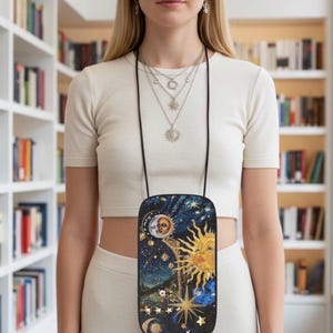 Pode incluir: Uma bolsa transversal preta para telefone com um design celestial com um sol, lua, estrelas e constelações. A bolsa é usada em volta do pescoço com um cordão preto. O fundo inclui uma estante.